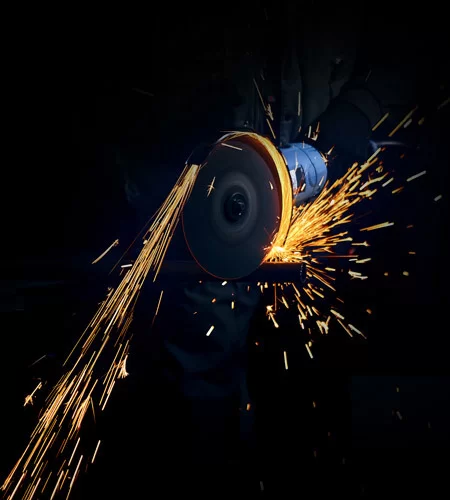 Cette image montre un ouvrier en train d'utiliser une meuleuse pour couper du métal, produisant une vive gerbe d'étincelles. Le travail est effectué dans un environnement sombre, accentuant la brillance et l'intensité des étincelles jaune-orange émanant de l'outil. La concentration sur l'action de la meuleuse met en évidence la puissance et la chaleur du processus de découpe. | Recyclage LGI